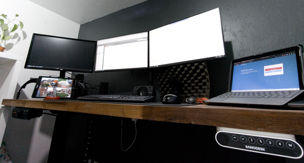 A dark wooden standing desk, with multiple screens a tablet, keyboard and mouse within a grey and white walled office